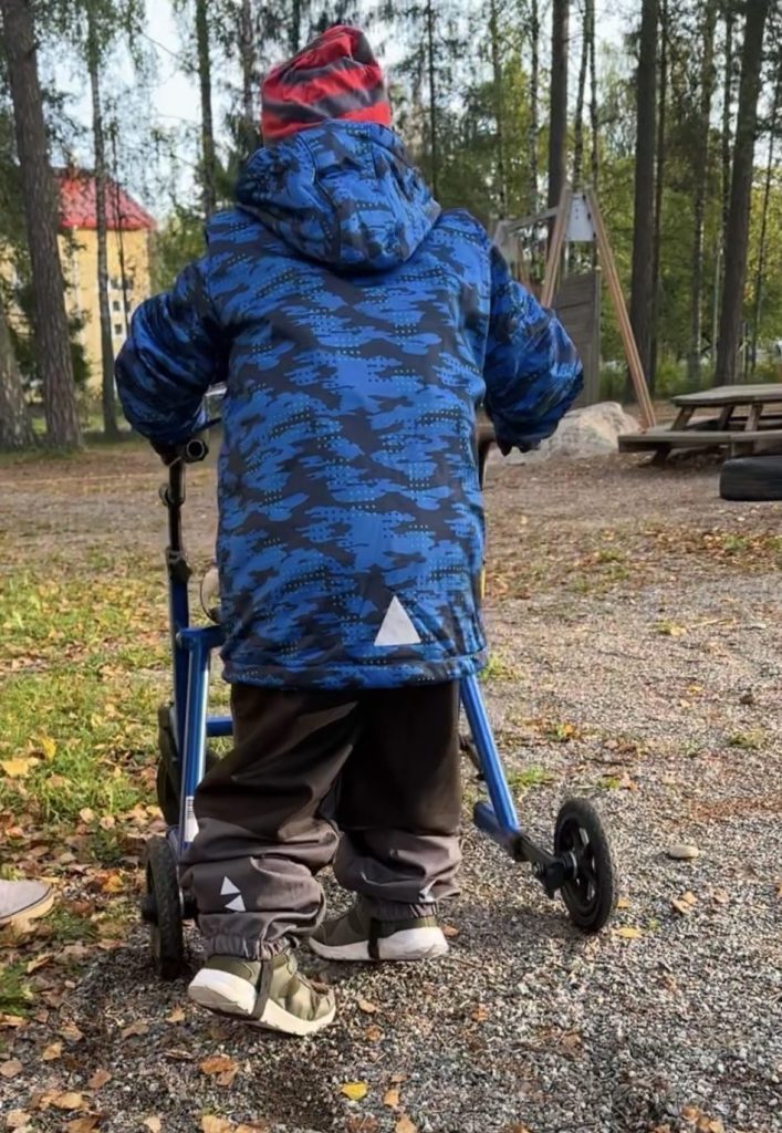 Lapsella on sininen takki ja punainen pipo. Hänet on kuvattu takaapäin kävelemässä sinisellä rollaattorilla syksyisellä pihalla.