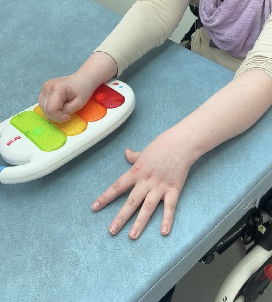 Kädet soittavat lasten lelupianoa. Oikea käsi on nyrkissä soittimen päällä, vasen avoinna vierellä. Soittimessa on eri värisiä painikkeita, joihin syttyy valot.