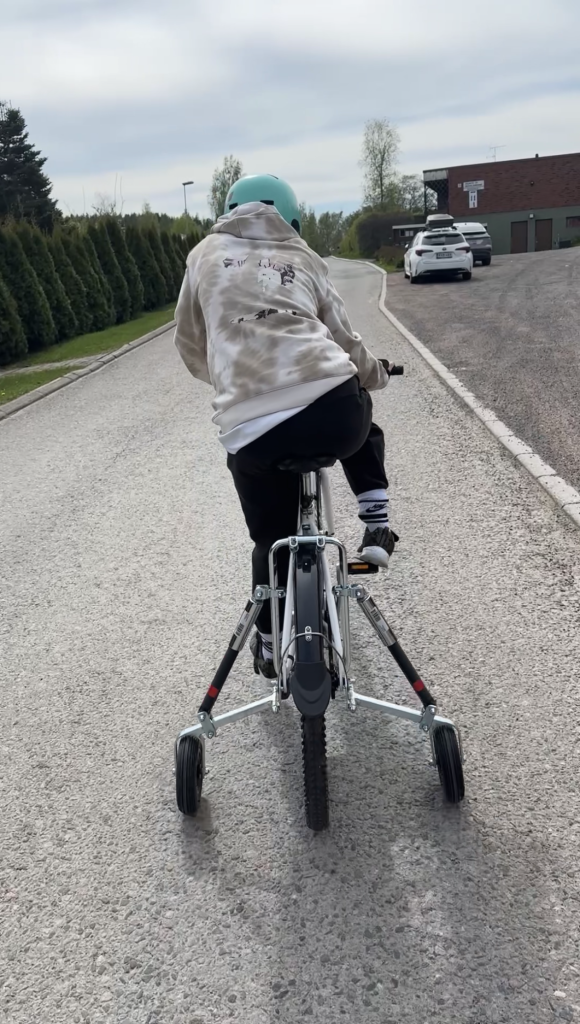 Lapsi kuvattuna takaa, polkemassa apupyörällistä pokupyörää. Lapsella on harmaa huppari ja turkoosi pyöräilykypärä.